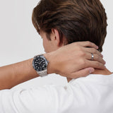 Ανδρικό ρολόι Daniel Wellington Iconic GMT DW00100919 φορεμένο με ασημί ατσάλινο μπρασελέ, μαύρο καντράν, γκρι λεπτομέρειες και μαύρη κεραμική στεφάνη.