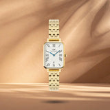 Γυναικείο ρολόι Daniel Wellington Quadro Roman Numerals DW00100861 με χρυσό μπρασελέ από ανοξείδωτο ατσάλι και λευκό καντράν με μπλε δείκτες μεγέθους 20x26mm.