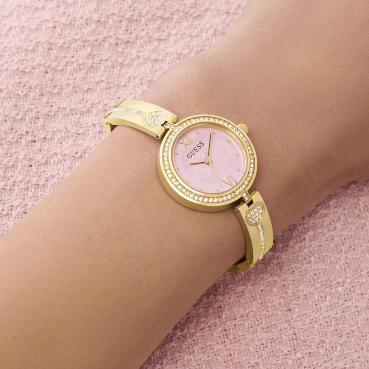 Γυναικείο ρολόι Guess Lovey GW0655L5 φορεμένο με χρυσό μπρασελέ, ροζ καντράν και διακόσμηση με ζιργκόν.
