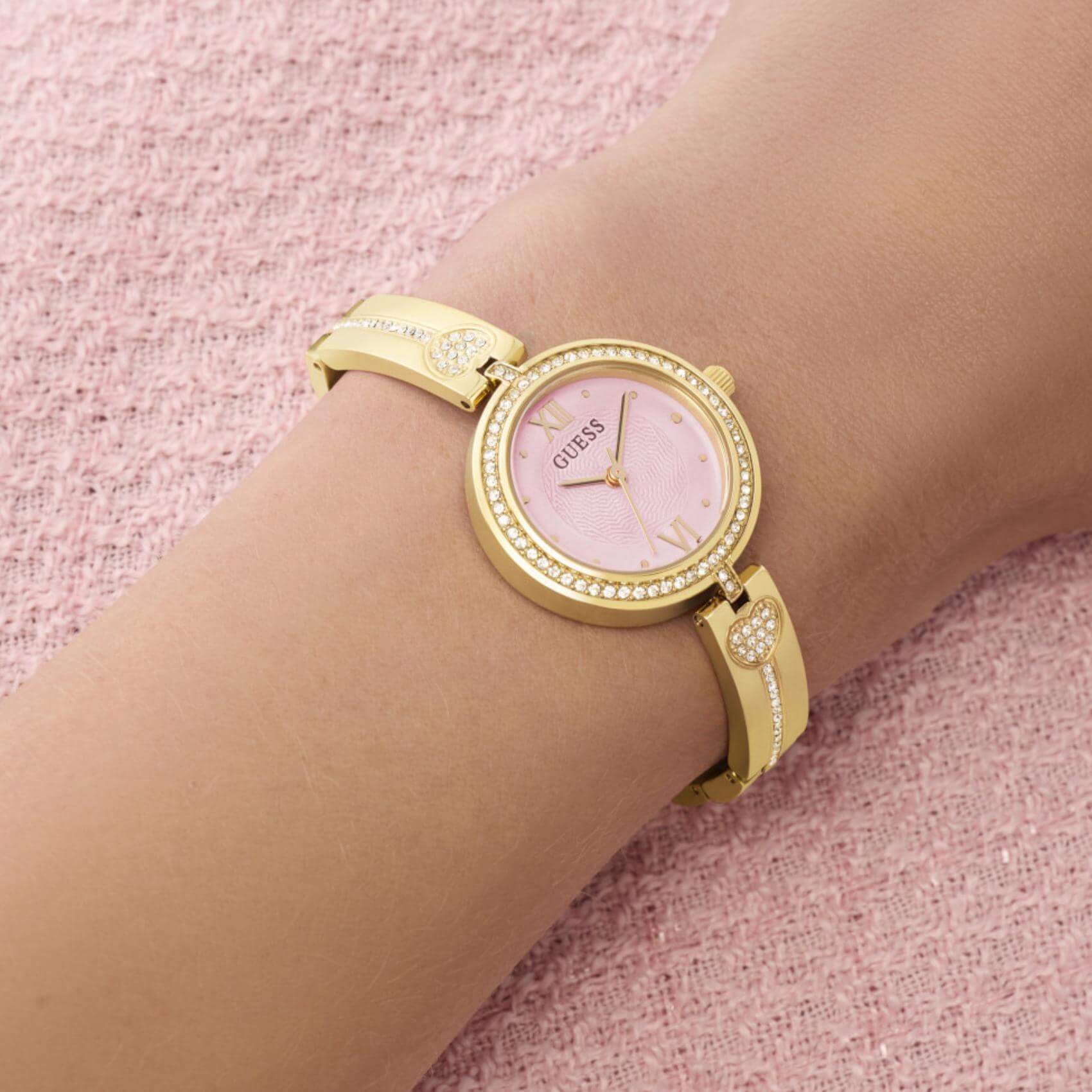 Γυναικείο ρολόι Guess Lovey GW0655L5 φορεμένο με χρυσό μπρασελέ, ροζ καντράν και διακόσμηση με ζιργκόν.