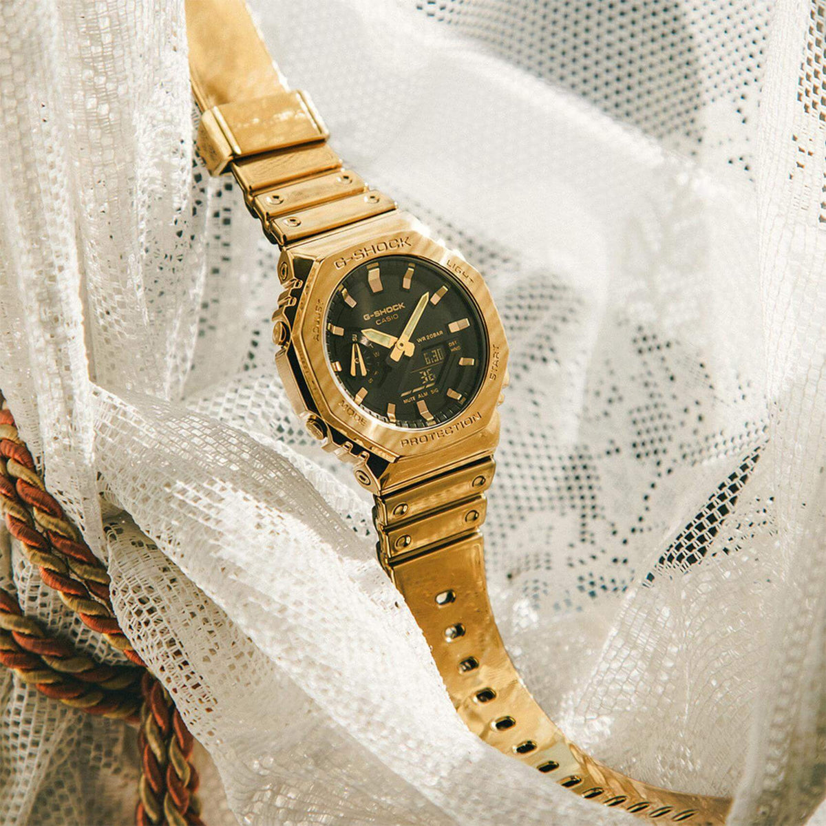 Ρολόι Casio G-Shock GM-2100YMG-9AER με χρυσό καουτσούκ λουράκι και οκτάγωνο μαύρο καντράν 45,4mm. με ημερομηνία-σε λευκό ύφασμα.