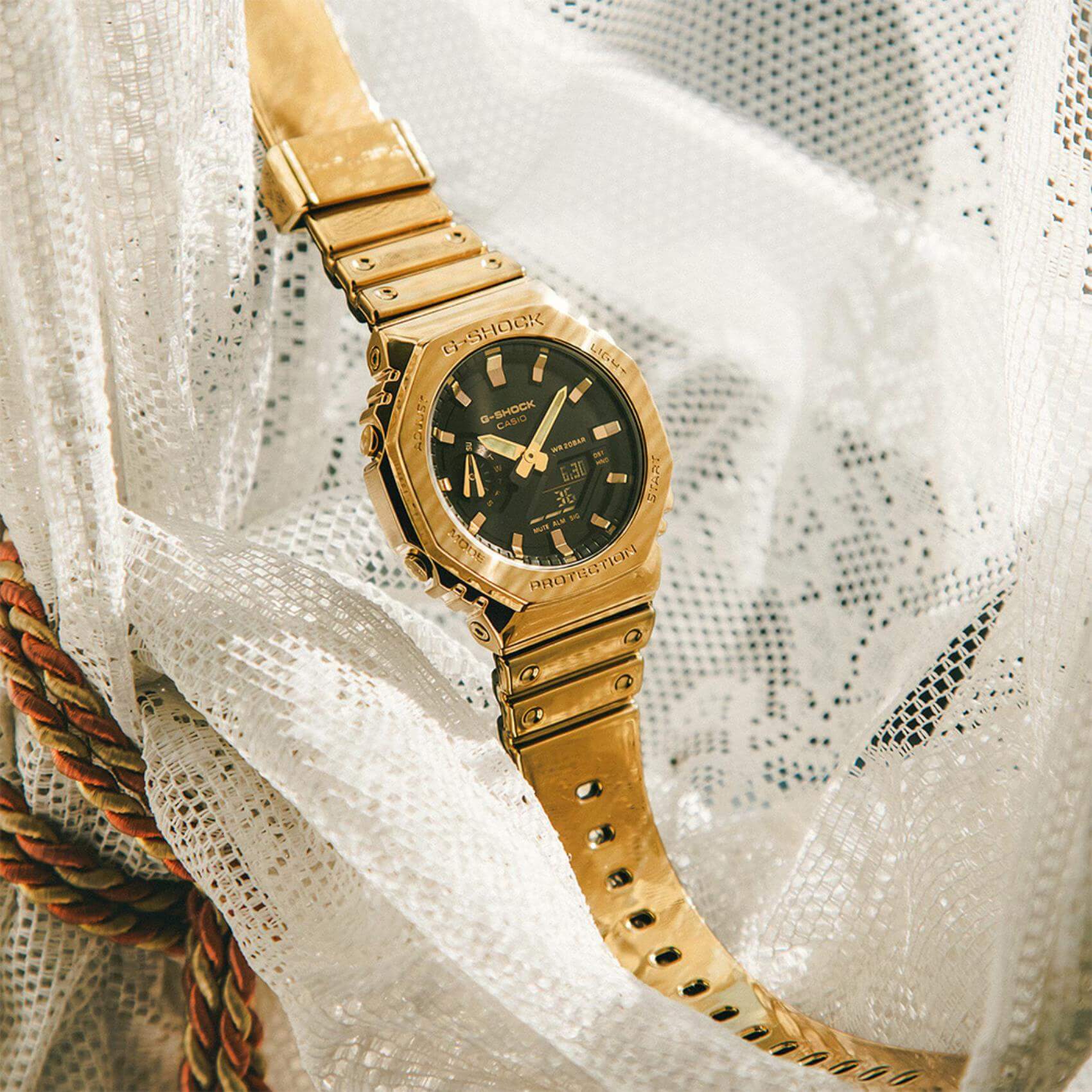 Ρολόι Casio G-Shock GM-2100YMG-9AER με χρυσό καουτσούκ λουράκι και οκτάγωνο μαύρο καντράν 45,4mm. με ημερομηνία-σε λευκό ύφασμα.