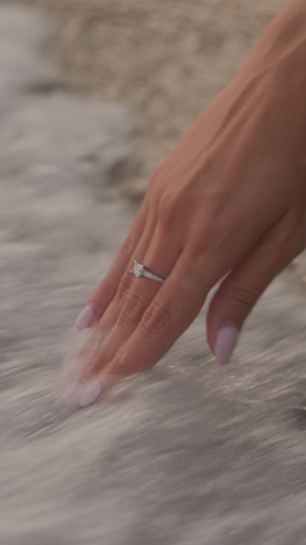 Λευκόχρυσο μονόπετρο δαχτυλίδι με κεντρικό διαμάντι, πλαϊνές πέτρες από διαμάντια και καστόνι με τέσσερα δόντια, φορεμένο σε γυναικείο χέρι.