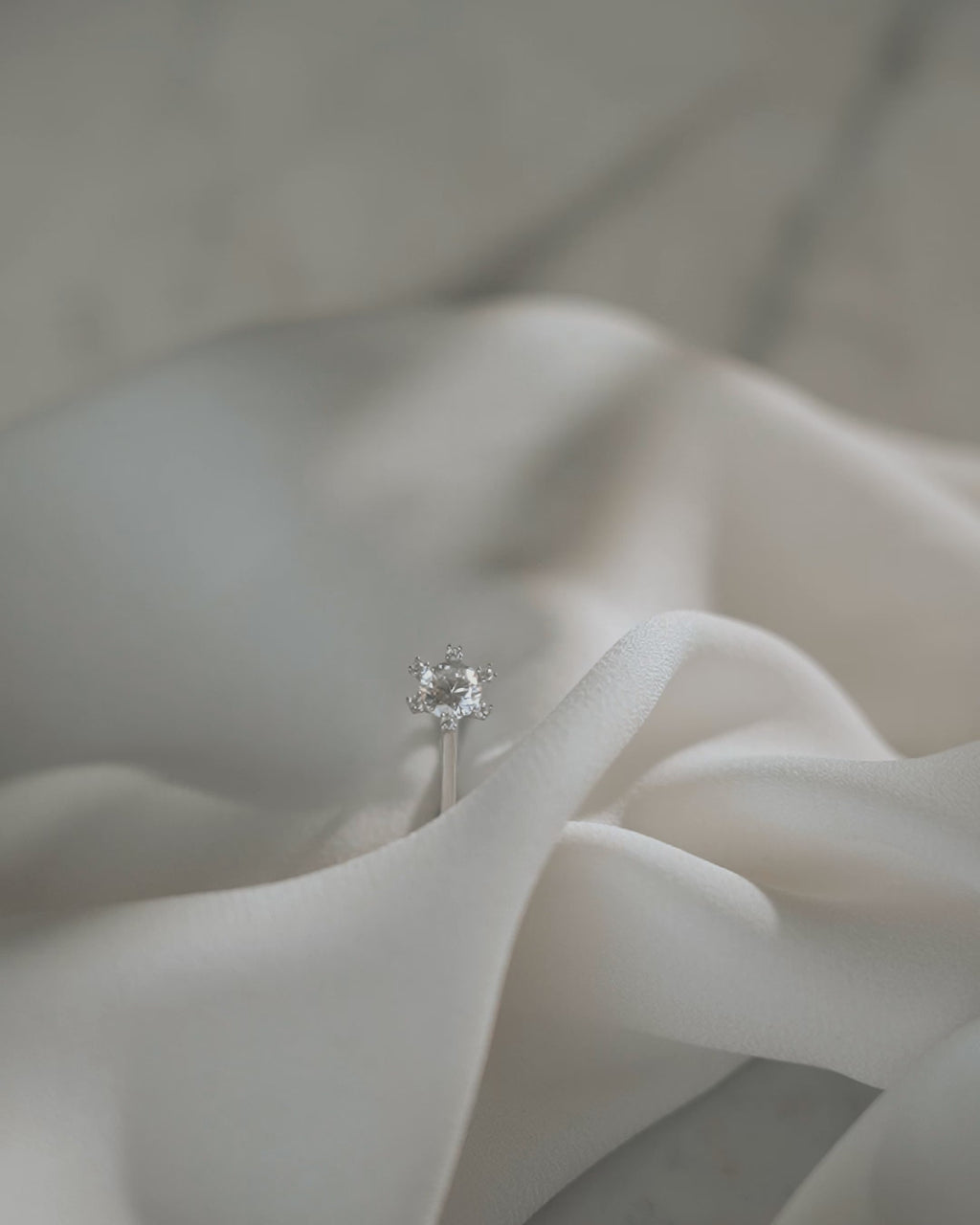 Μονόπετρο δαχτυλίδι γάμου με κεντρικό διαμάντι και πέτρες brilliant περιμετρικά κατασκευασμένο από λευκόχρυσο, επάνω σε λευκό μετάξι.