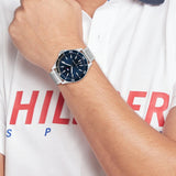 Ανδρικό ρολόι Tommy Hilfiger Logan 1792012, με μπρασελέ σε ασημί χρώμα από ανοξείδωτο ατσάλι και μπλε καντράν φορεμένο σε ανδρικό χέρι.