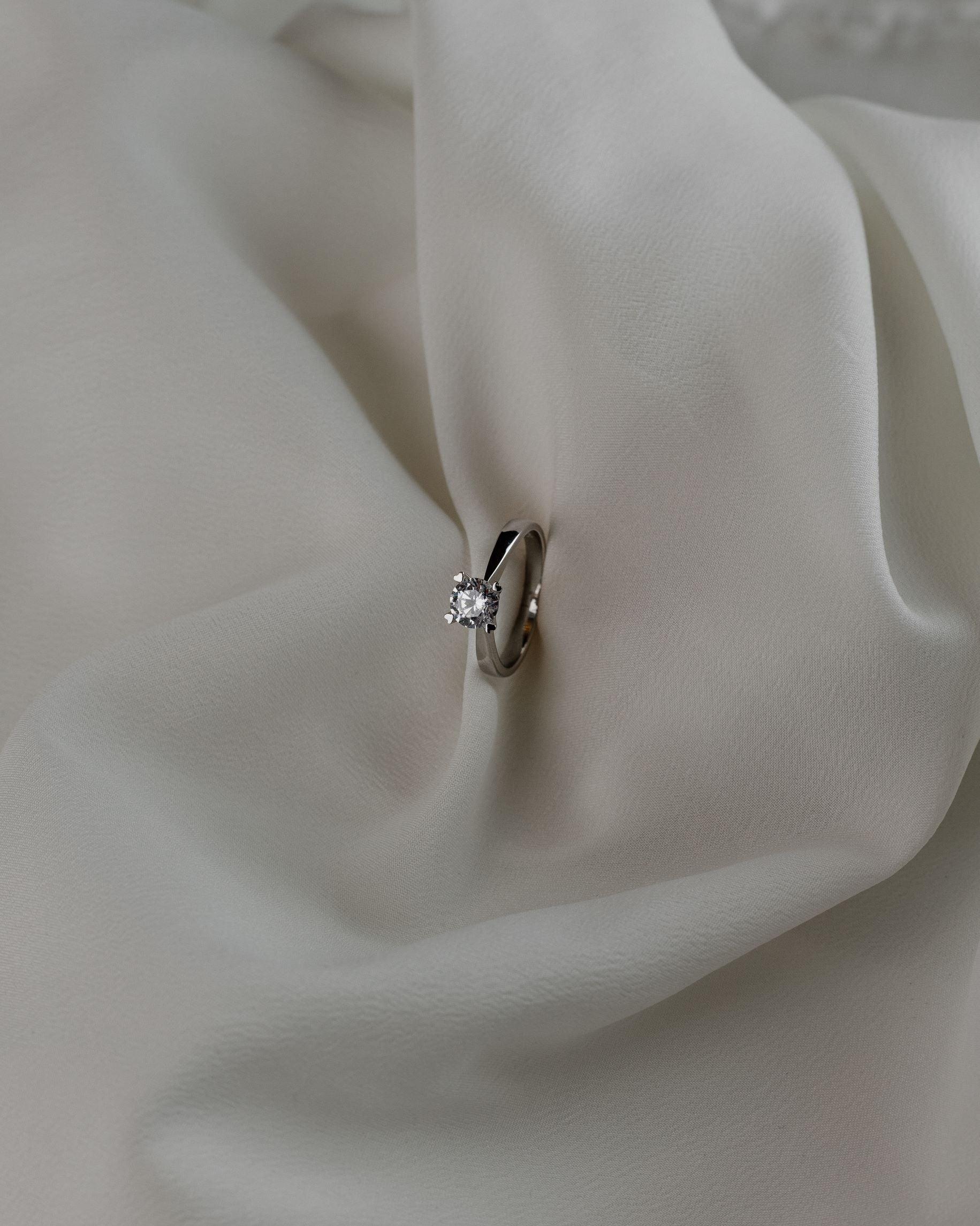 Λευκόχρυσο μονόπετρο με διαμάντι, με καστόνι σε σχήμα "V" και λεπτομέρειες σε σχήμα καρδιάς, επάνω σε λευκό μετάξι.
