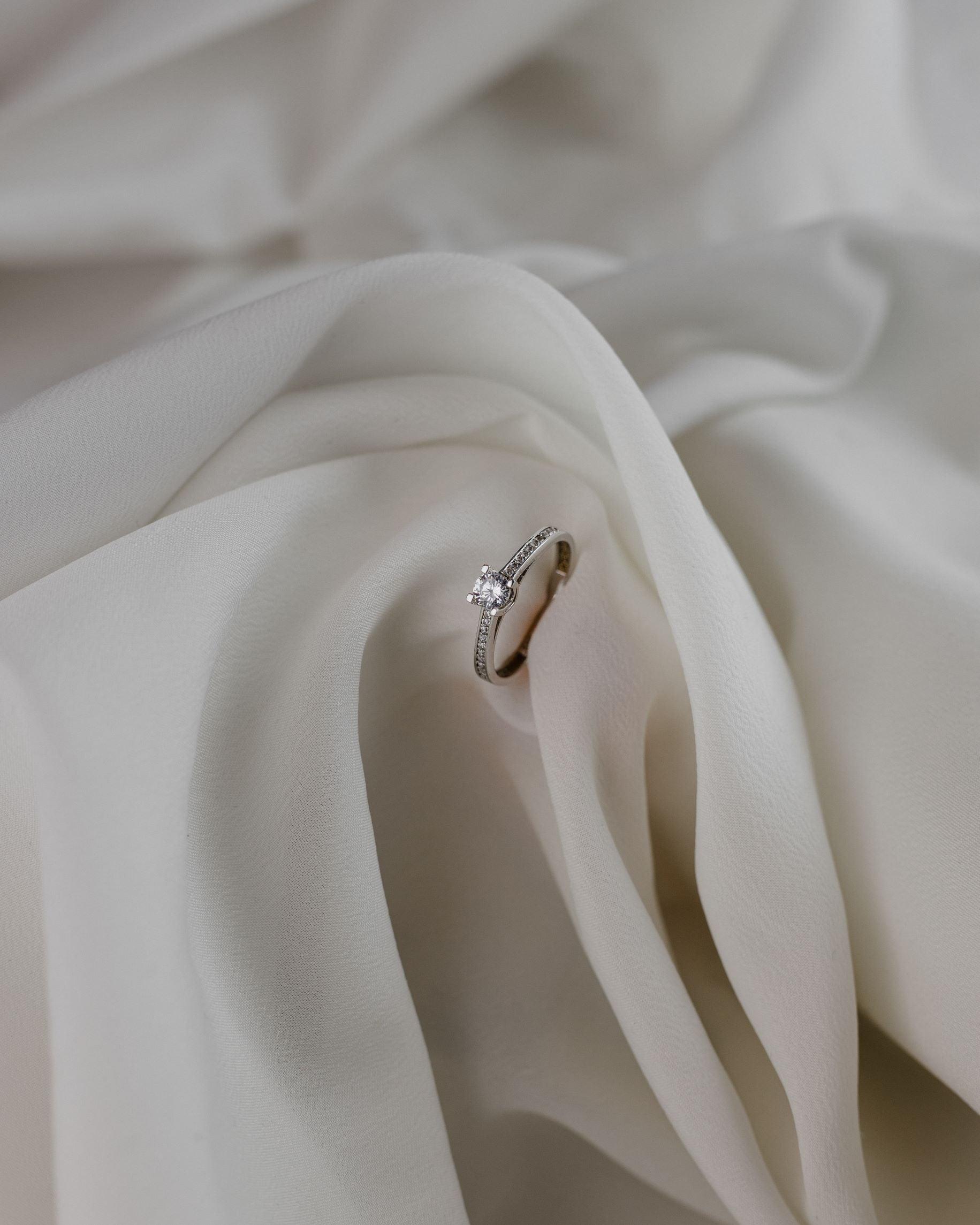 Λευκόχρυσο μονόπετρο με διαμάντι, καστόνι σε σχήμα "V" και πλαϊνές πέτρες, επάνω σε λευκό ύφασμα. 