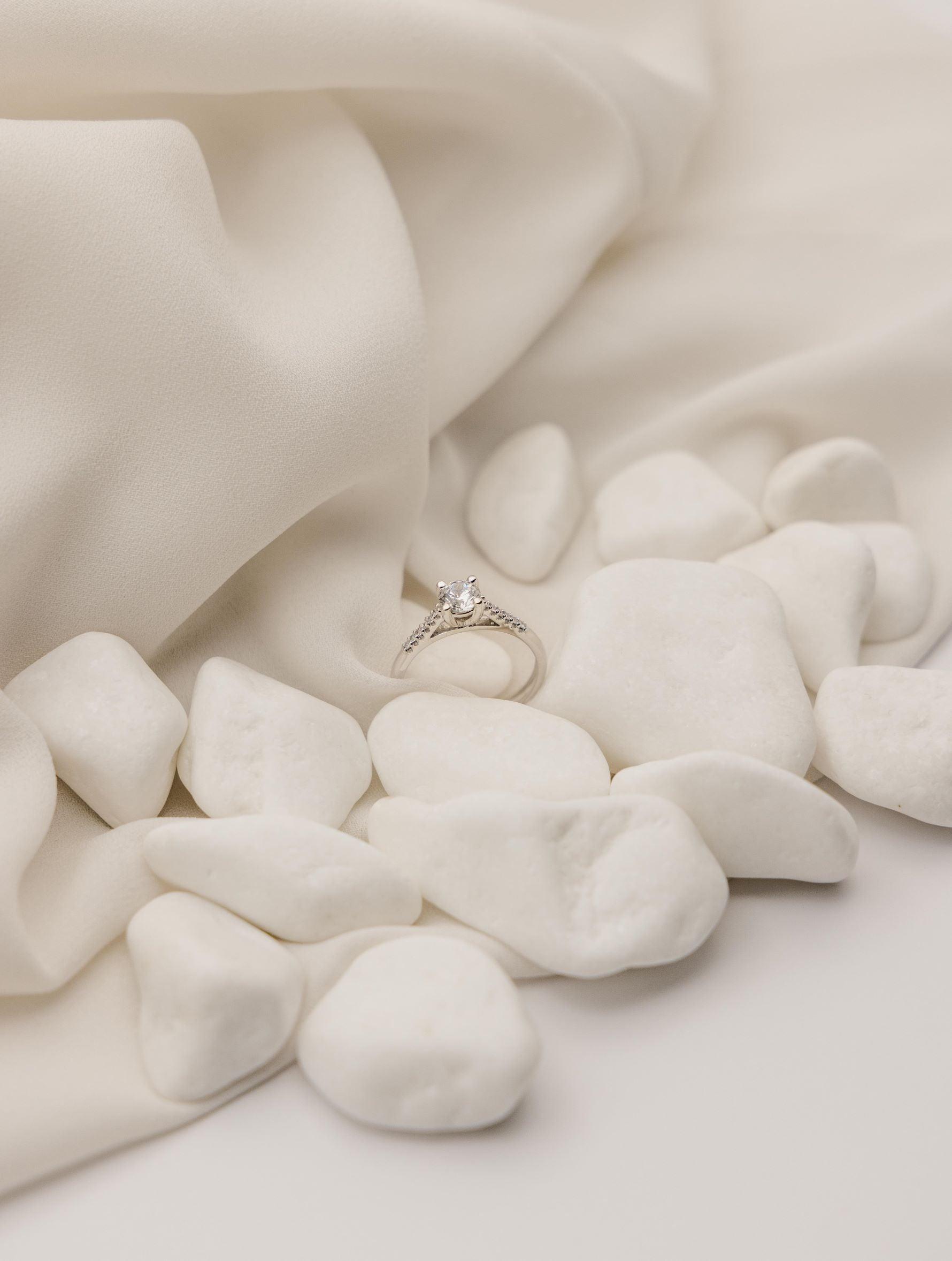 Λευκόχρυσο μονόπετρο δαχτυλίδι με διαμάντι και πλαίνές πέτρες από μικρότερα διαμάντια, επάνω σε λευκά βότσαλα.