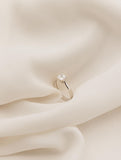 Λευκόχρυσο μονόπετρο δαχτυλίδι με διαμάντι και καστόνι σε σχήμα "V", επάνω σε λευκό ύφασμα.