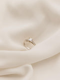 Λευκόχρυσο μονόπετρο δαχτυλίδι με κεντρικό διαμάντι, πλαϊνές πέτρες από διαμάντια και καστόνι με τέσσερα δόντια, επάνω σε λευκό μετάξι.
