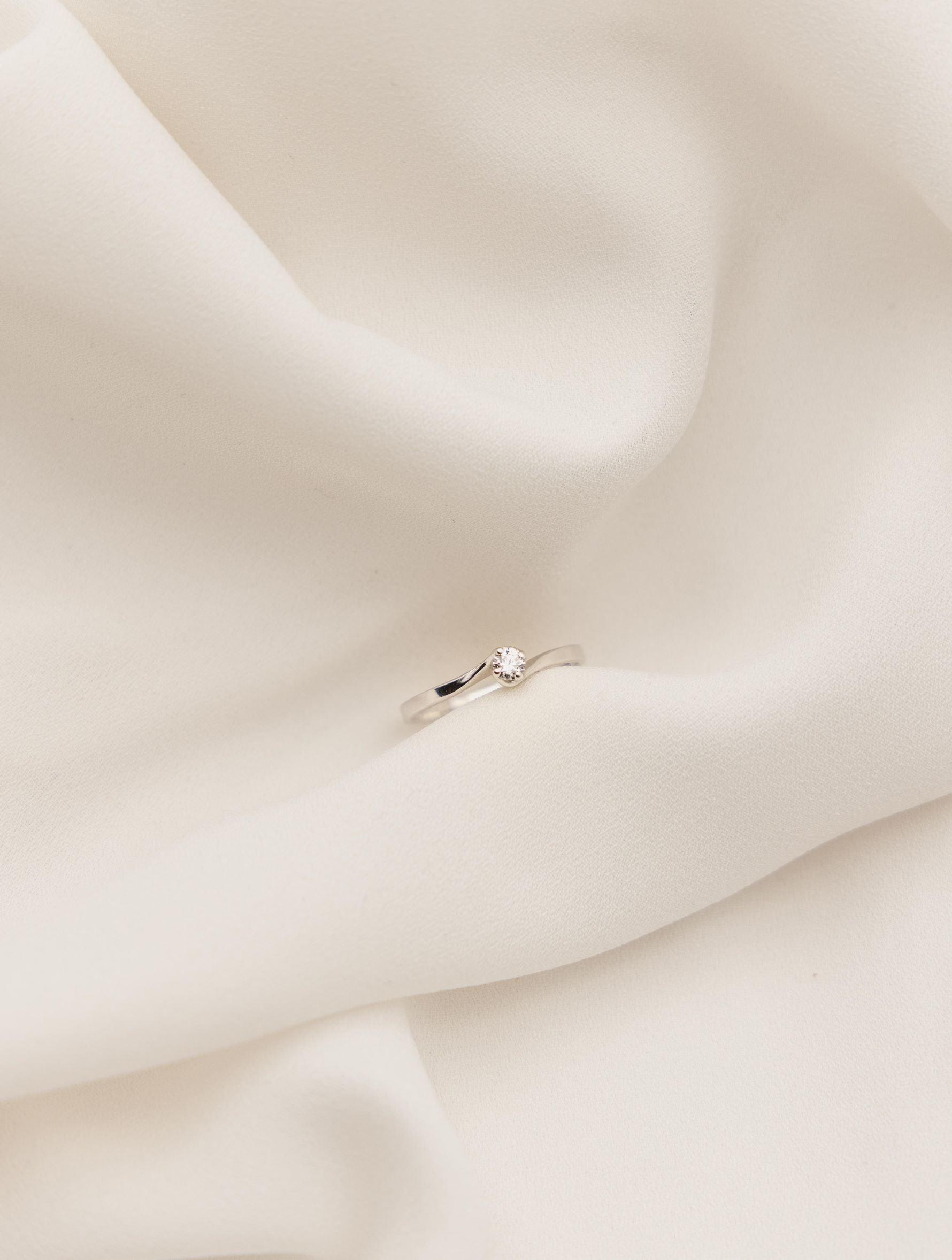 Λευκόχρυσο μονόπετρο δαχτυλίδι με διαμάντι και καστόνι που αγκαλιάζει την πέτρα σε σχήμα φλόγας, επάνω σε λευκό μετάξι.