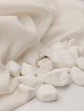 Λευκόχρυσο μονόπετρο δαχτυλίδι με διαμάντι και πλαίνές πέτρες από μικρότερα διαμάντια, επάνω σε λευκό μετάξι και βότσαλα.