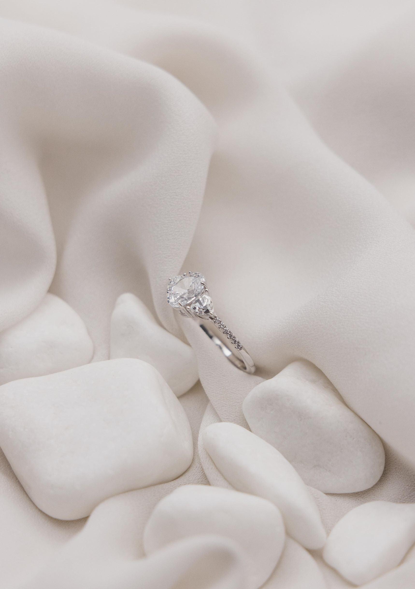 Μονόπετρο δαχτυλίδι με οβάλ κεντρικό διαμάντι και πλαίνές πέτρες από μικρότερα διαμάντια, κατασκευασμένο από λευκόχρυσο, επάνω σε λευκό μετάξι.