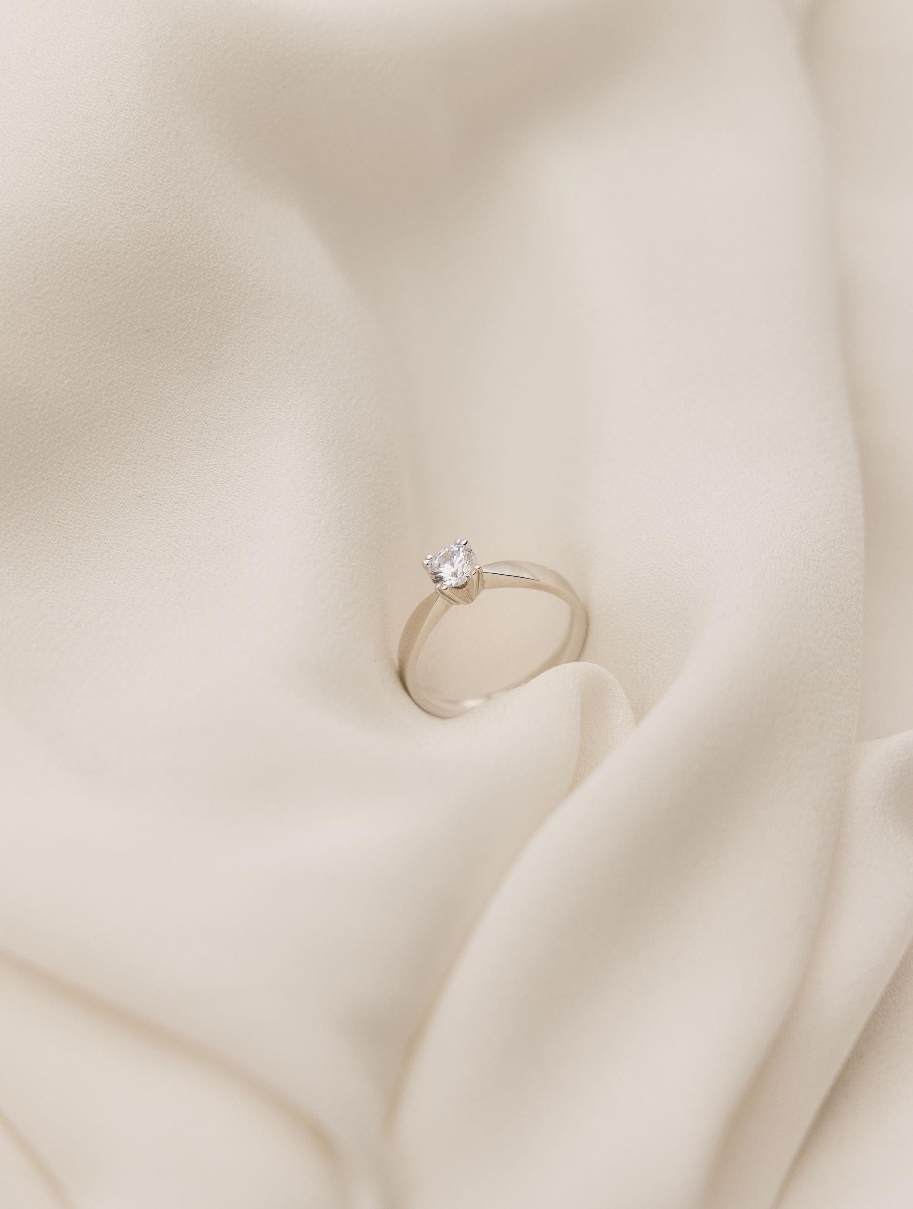 Λευκόχρυσο μονόπετρο δαχτυλίδι με διαμάντι και καστόνι σε σχήμα "V", επάνω σε λευκό μετάξι.