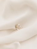 Λευκόχρυσο μονόπετρο δαχτυλίδι με διαμάντι και πλαίνές πέτρες από μικρότερα διαμάντια, επάνω σε λευκό μετάξι.