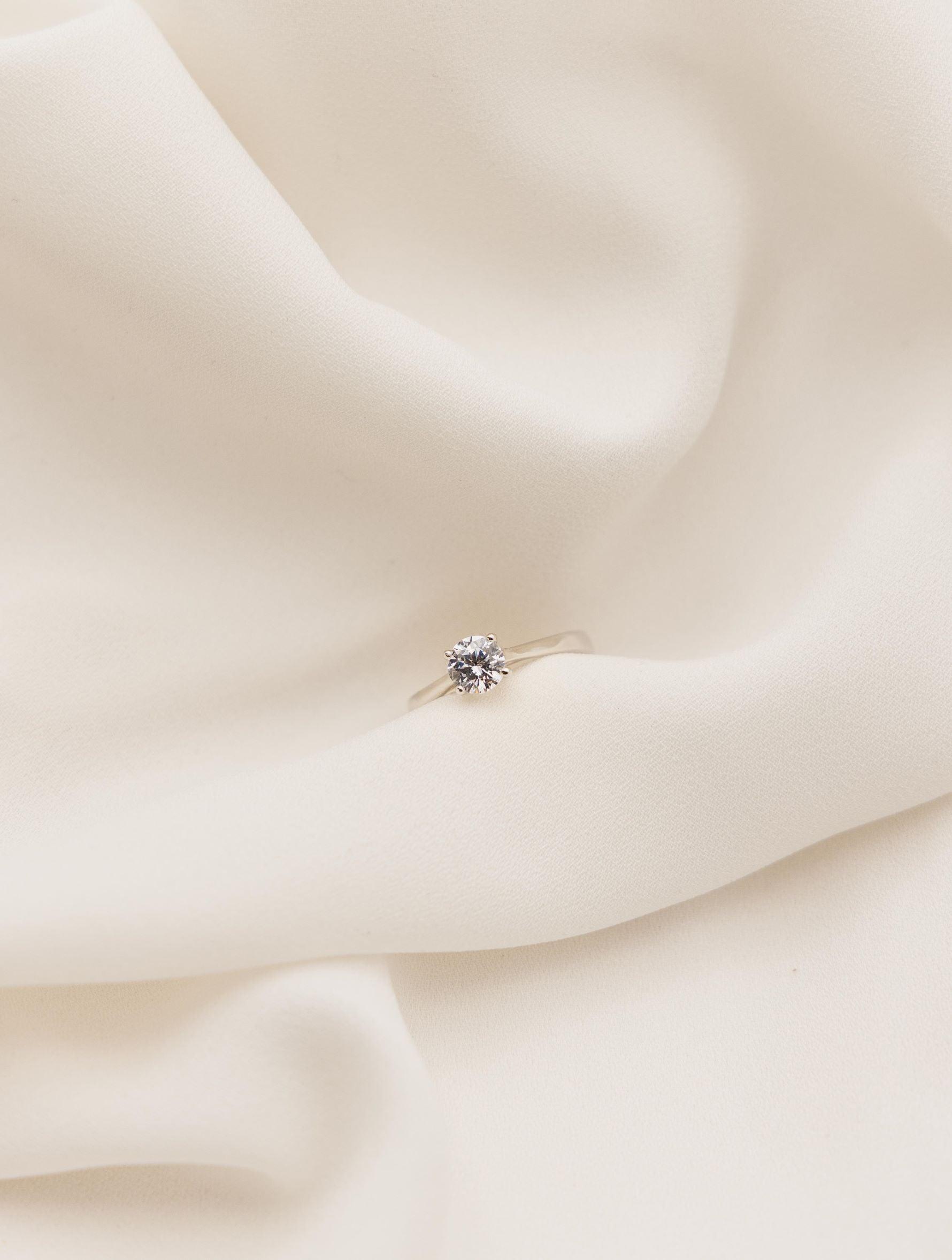 Λευκόχρυσο μονόπετρο δαχτυλίδι με διαμάντι και καστόνι που αγκαλιάζει την πέτρα με τέσσερα δόντια, επάνω σε λευκό μετάξι.