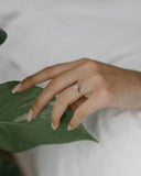 Μονόπετρο δαχτυλίδι γάμου με κεντρικό διαμάντι και πέτρες brilliant περιμετρικά κατασκευασμένο από λευκόχρυσο, φορεμένο σε γυναικείο χέρι.