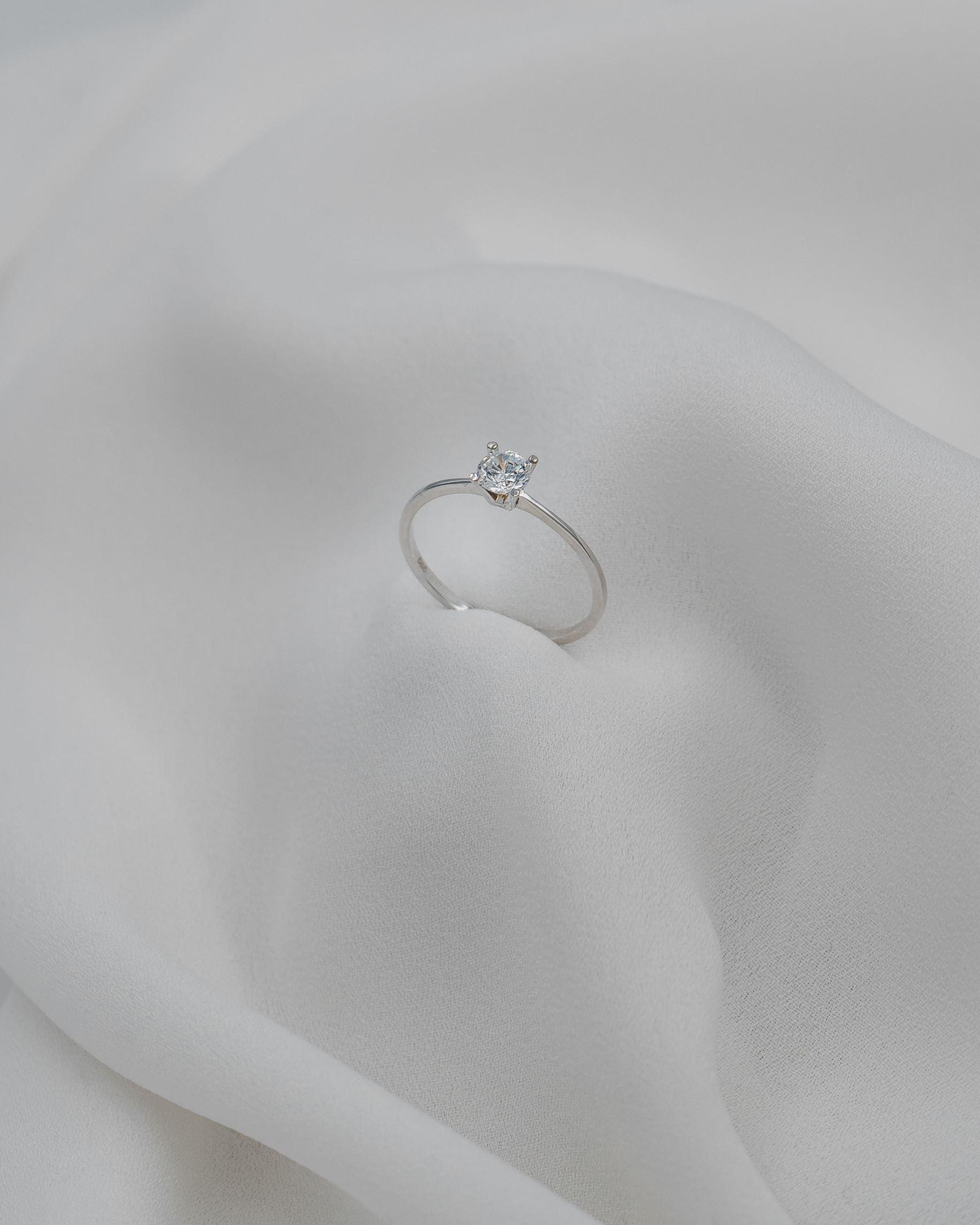 Μονόπετρο δαχτυλίδι γάμου με κεντρικό διαμάντι σε καστόνι σχήματος "V" κατασκευασμένο από λευκόχρυσο, ακουμπισμένο σε λευκό μετάξι.