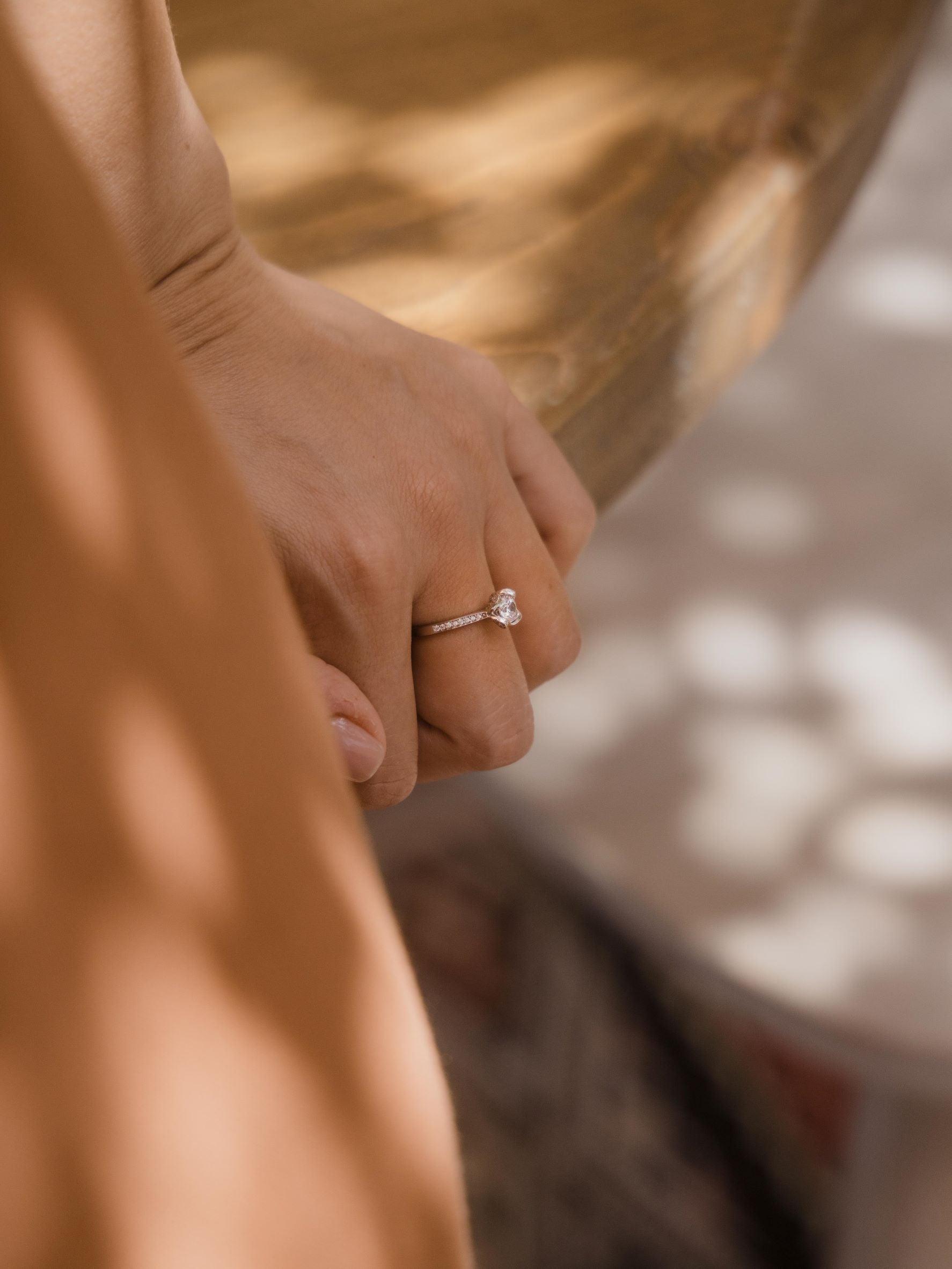 Μονόπετρο δαχτυλίδι με διαμάντι και πλαϊνές πέτρες από μικρότερα διαμάντια, κατασκευασμένο από λευκόχρυσο με καστόνι σε σχήμα λουλουδιού, φορεμένο σε γυναικείο χέρι.