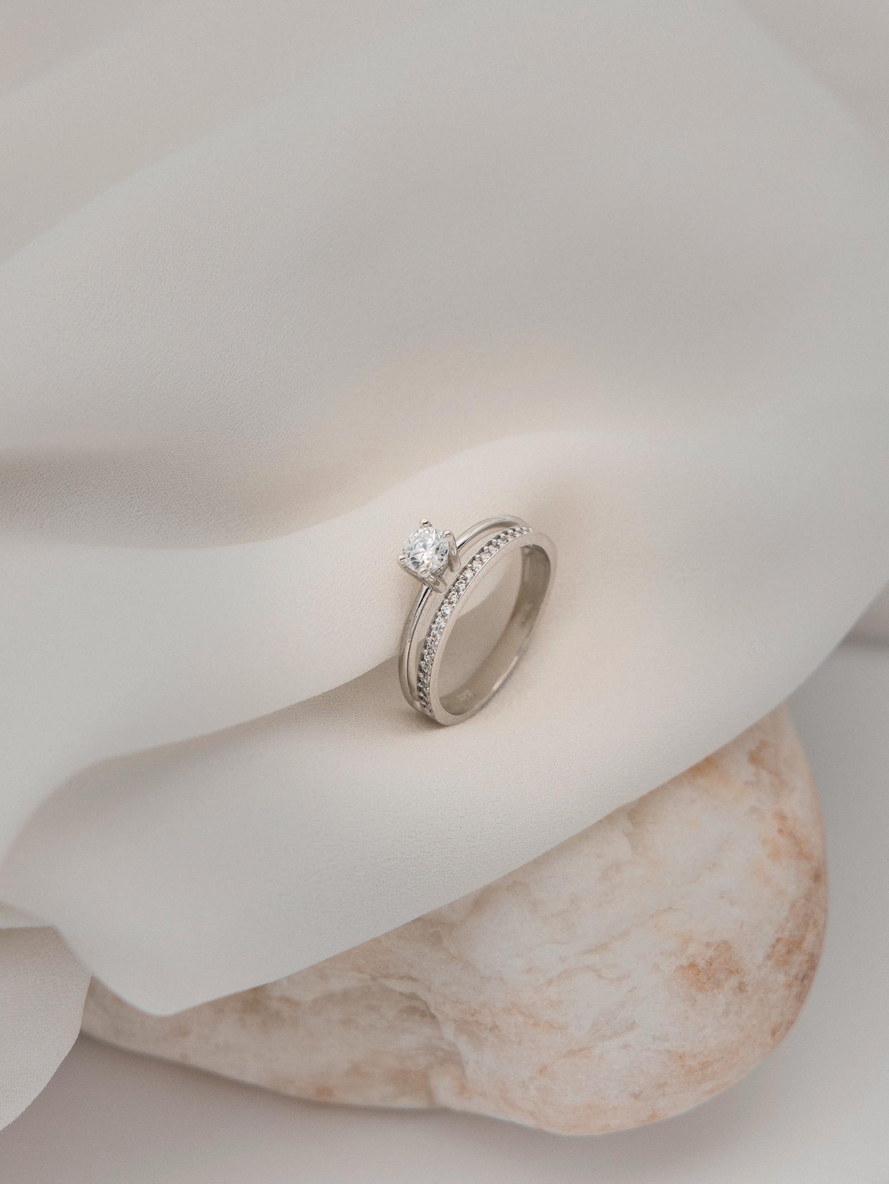 Μονόπετρο δαχτυλίδι γάμου με διαμάντι, που συνδυάζει ένα δαχτυλίδι σειρέ με διαμάντια δίνοντας την αίσθηση διπλού δαχτυλιδιού, κατασκευασμένο από λευκόχρυσο, τοποθετημένο επάνω σε λευκό ύφασμα. 