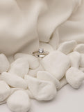 Ένα μονόπετρο δαχτυλίδι με διαμάντι κατασκευασμένο από λευκόχρυσο, με καστόνι από τέσσερα δόντια, τοποθετημένο επάνω σε λευκό ύφασμα με βότσαλα.