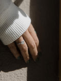 Μονόπετρο δαχτυλίδι με κεντρικό διαμάντι round cut, πλαίνές πέτρες από μικρότερα διαμάντια και στρογγυλό καστόνι σε σχήμα κορώνας κατασκευασμένο από λευκόχρυσο φορεμένο σε γυναικείο χέρι.