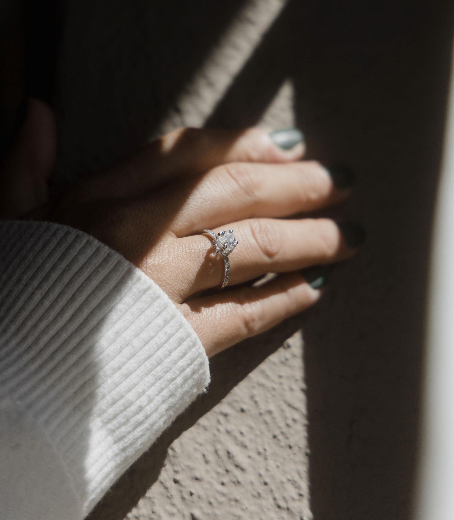 Μονόπετρο δαχτυλίδι με κεντρικό διαμάντι round cut, πλαίνές πέτρες από μικρότερα διαμάντια και στρογγυλό καστόνι σε σχήμα κορώνας κατασκευασμένο από λευκόχρυσο φορεμένο σε γυναικείο χέρι.