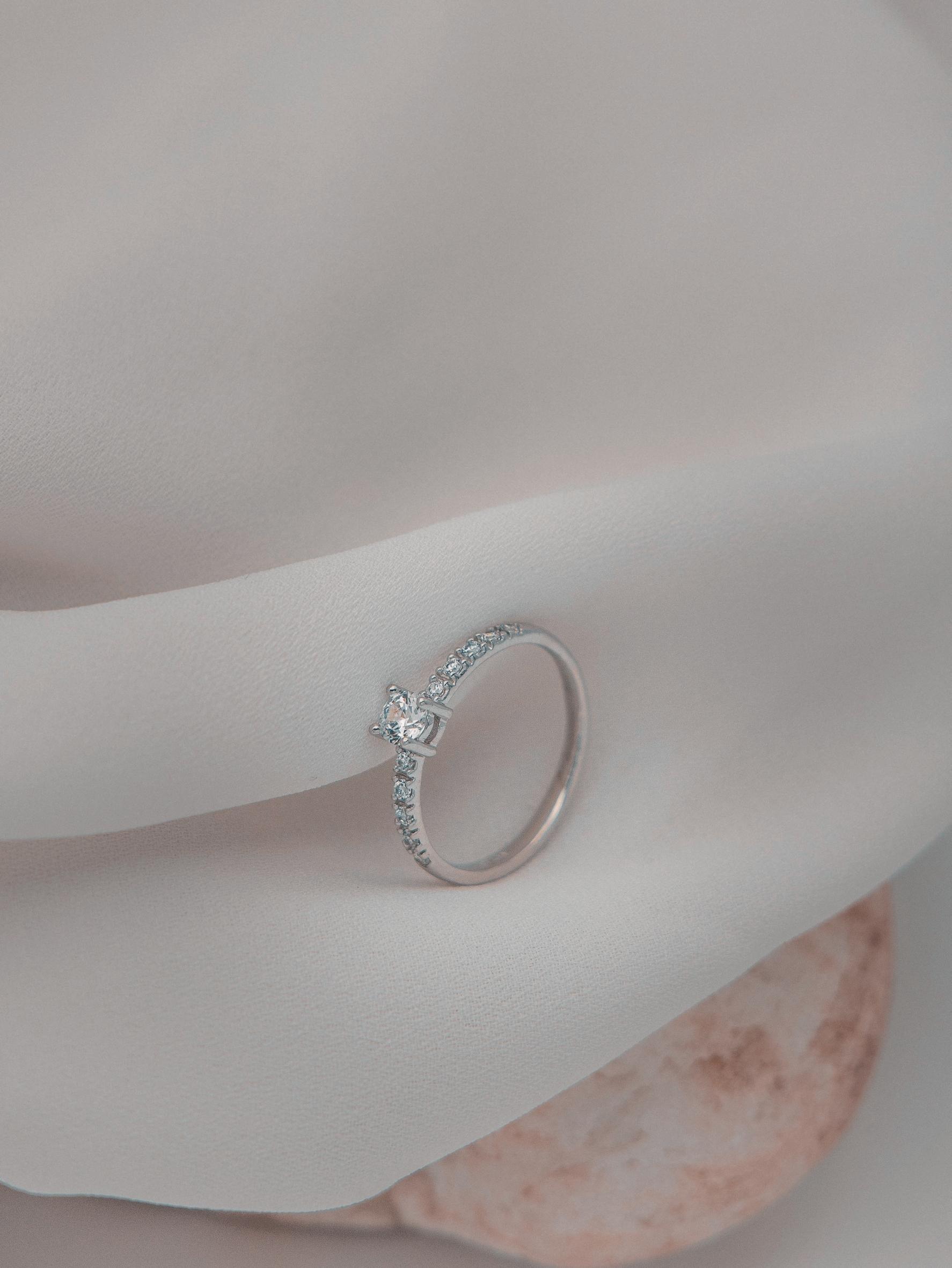 Μονόπετρο δαχτυλίδι με διαμάντι, κατασκευασμένο από λευκόχρυσο. πλαϊνές πέτρες από μικρότερα διαμάντια και καστόνι με τέσσερα δόντια, επάνω σε λευκό ύφασμα. 