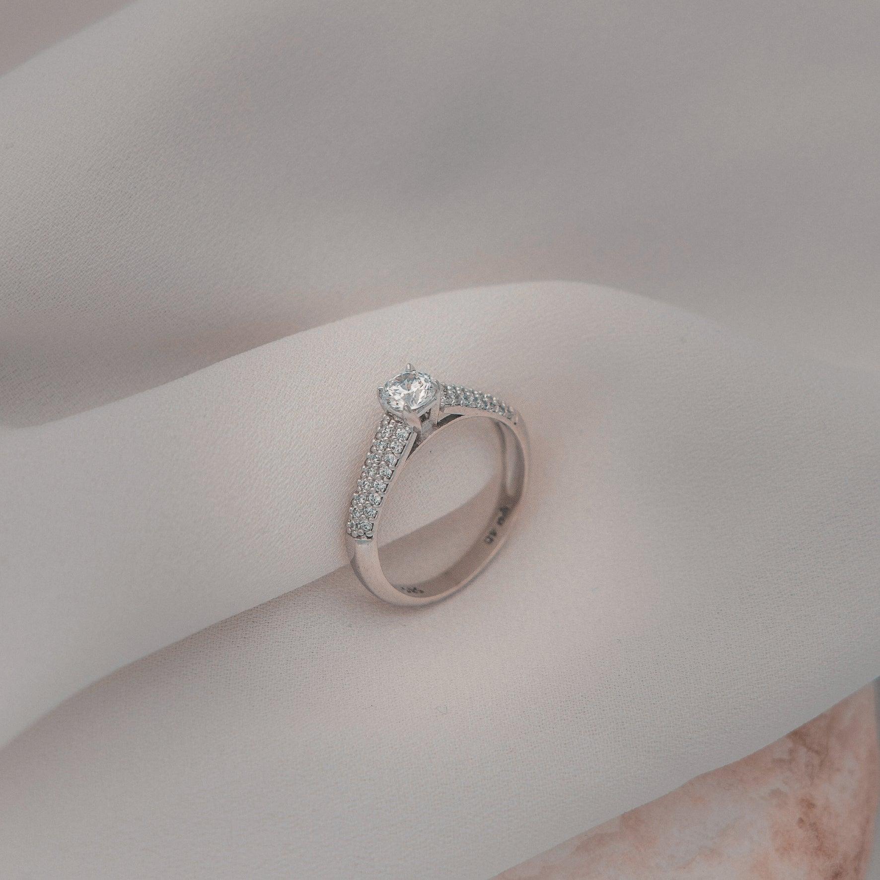 Μονόπετρο δαχτυλίδι με κεντρικό διαμάντι round cut και πλαϊνές πέτρες από μικρότερα διαμάντια, κατασκευασμένο από λευκόχρυσο. 