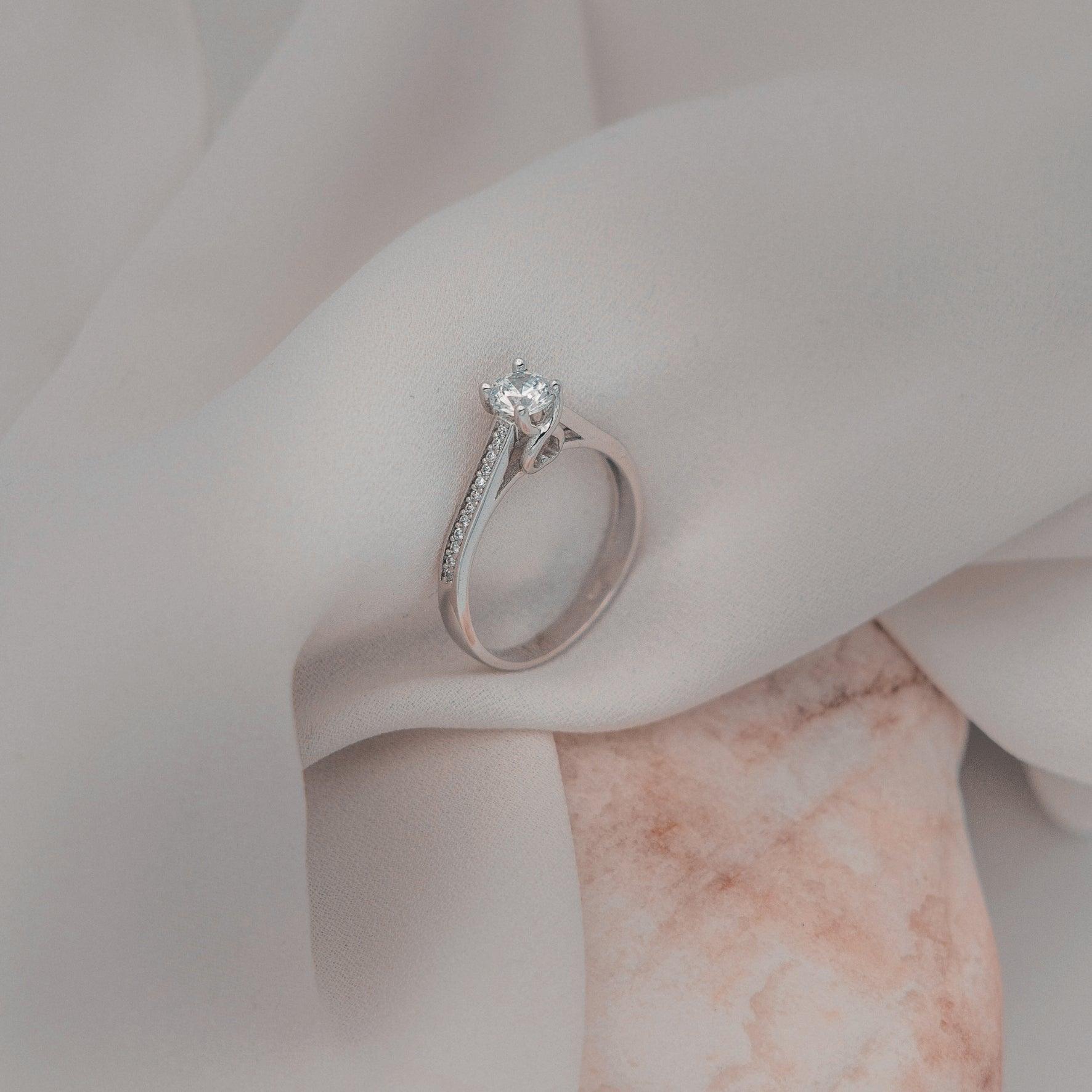 Μονόπετρο δαχτυλίδι με κεντρικό διαμάντι round cut και πλαϊνές πέτρες από μικρότερα διαμάντια, κατασκευασμένο από λευκόχρυσο.
